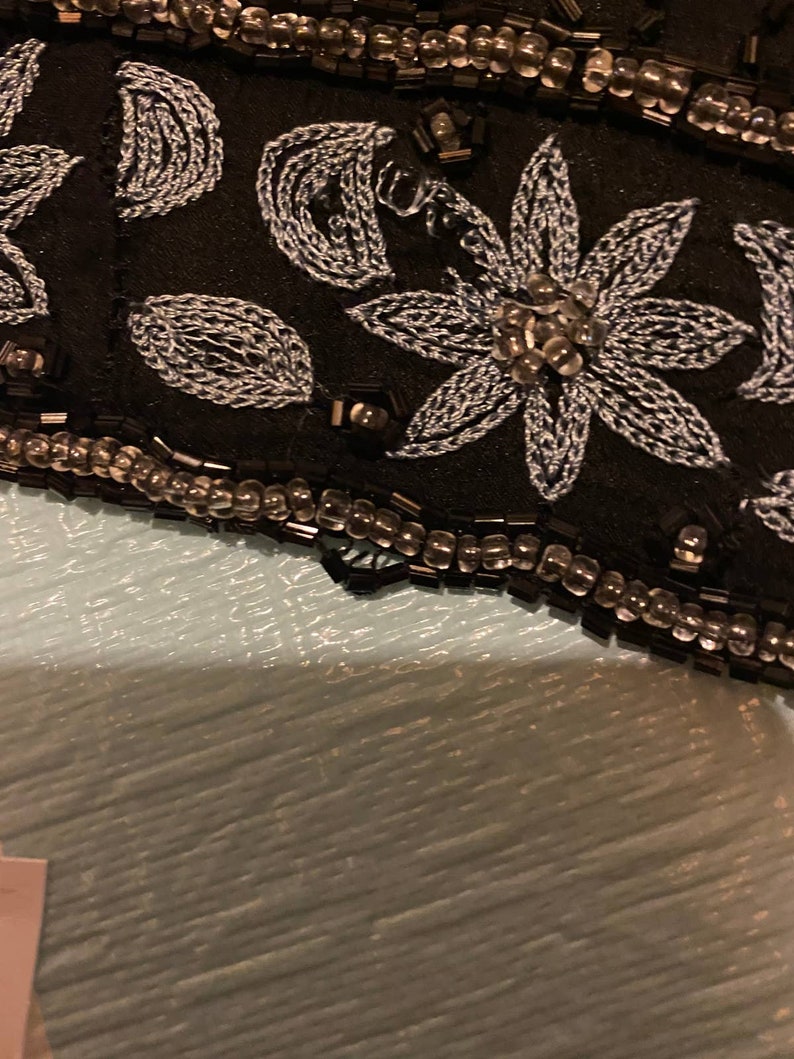 May include: Close-up of a black fabric with silver embroidery of flowers and leaves. The fabric is trimmed with a row of small, clear beads.