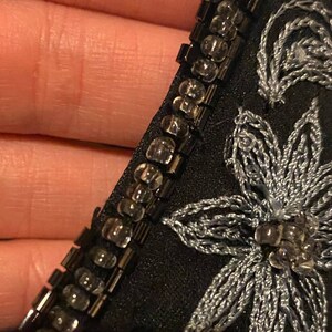 May include: Close-up of a black fabric with silver embroidery and a beaded trim. The trim is made of small, clear beads.