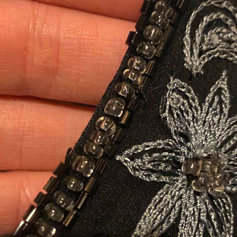 May include: Close-up of a black fabric with silver embroidery and a beaded trim. The trim is made of small, clear beads.