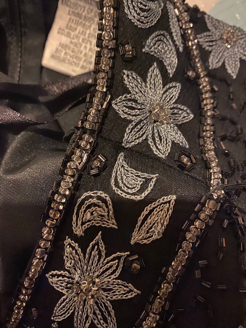 May include: Close-up of a black fabric with silver and black beading and embroidery. The embroidery features a floral design with leaves and flowers.