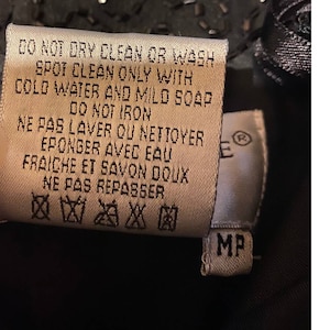 May include: A white care tag with black text detailing the care instructions for the garment. The tag reads "DO NOT DRY CLEAN OR WASH SPOT CLEAN ONLY WITH COLD WATER AND MILD SOAP DO NOT IRON NE PAS LAVER OU NETTOYER EPONGER AVEC EAU FRAICHE ET SAVON DOUX NE PAS REPASSER" and includes symbols for hand washing, not bleaching, not tumble drying, not dry cleaning, and not ironing. The tag also has a small white tag with the letter "E" and the symbol for registered trademark, and a white tag with the letters "MP".