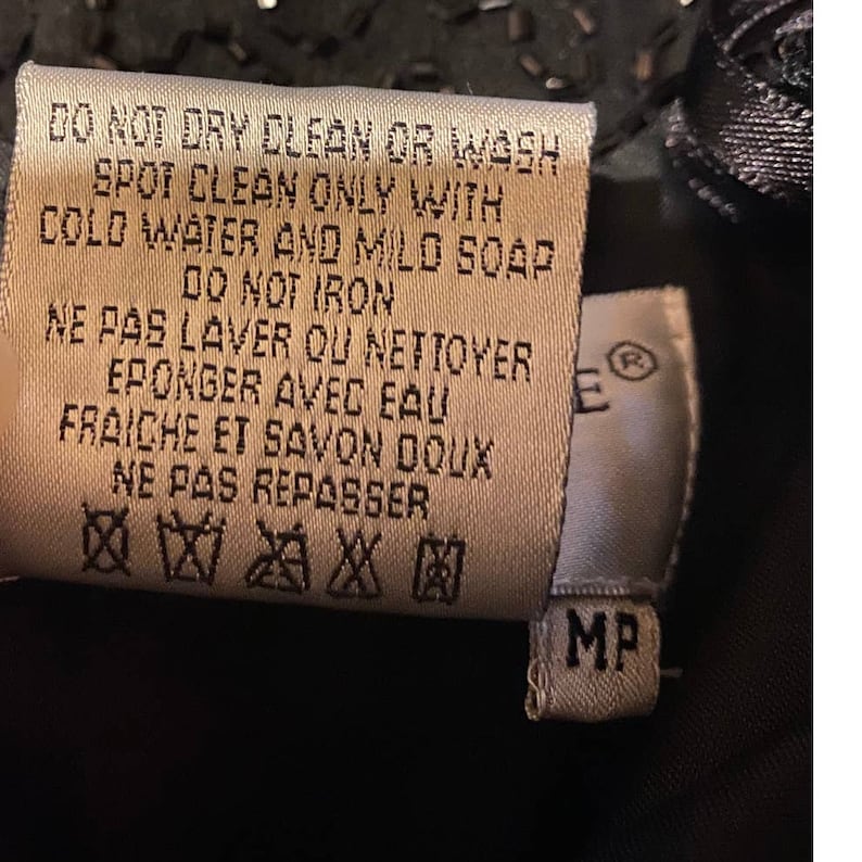 May include: A white care tag with black text detailing the care instructions for the garment. The tag reads "DO NOT DRY CLEAN OR WASH SPOT CLEAN ONLY WITH COLD WATER AND MILD SOAP DO NOT IRON NE PAS LAVER OU NETTOYER EPONGER AVEC EAU FRAICHE ET SAVON DOUX NE PAS REPASSER" and includes symbols for hand washing, not bleaching, not tumble drying, not dry cleaning, and not ironing. The tag also has a small white tag with the letter "E" and the symbol for registered trademark, and a white tag with the letters "MP".