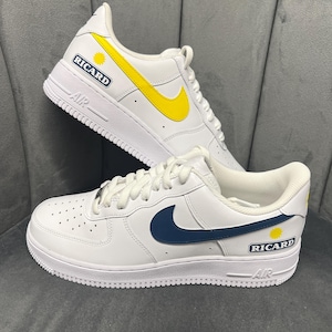 Peut inclure: Baskets en cuir blanc avec des détails jaunes et bleu marine. Les baskets présentent le mot "RICARD" et un motif de soleil jaune sur le côté. Le logo Nike est bleu marine. Les semelles sont blanches.