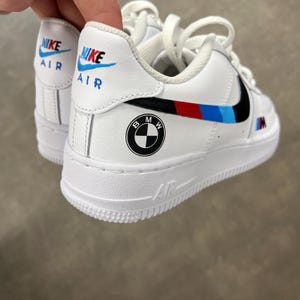 Può includere: Sneaker bianche con logo Nike Air sulla linguetta del tallone in blu e nero. Il lato mostra un logo BMW, uno swoosh a righe nere, rosse e azzurre e un piccolo logo M. La suola in gomma bianca presenta la scritta Air impressa sul lato.