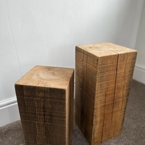May include: Two square wooden stools, one taller than the other, made of rough-hewn wood with a natural finish. The stools are sitting on a brown carpet.