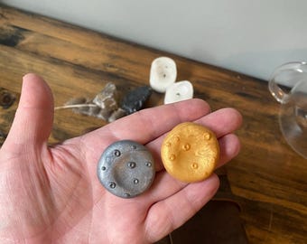 May include: Two round, decorative objects are held in a hand. One is silver with small, dark indentations, and the other is gold with a textured surface. The objects are on a wooden surface.