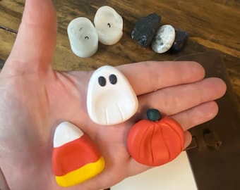 May include: A hand holds several Halloween-themed clay figures. There is a white ghost with black eyes, a candy corn, and a small orange pumpkin. Also present are two white candle-shaped figures and several small black and white stones.