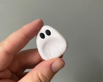 May include: A small, white ghost-shaped figurine with two black dot eyes. The figurine is smooth and rounded, with subtle lines suggesting a draped form. The object is held in a hand against a neutral gray background.