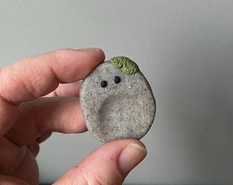 May include: A small, gray stone figurine with black dot eyes and a green leaf-like detail on top. The stone has a speckled texture and a smooth, rounded shape. The figurine is held in a person's hand.