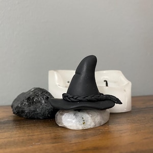 Puede incluir: Un sombrero de bruja negro con una banda trenzada se sienta sobre un cristal blanco. El sombrero está hecho de arcilla polimérica y tiene una punta puntiaguda.