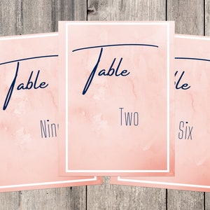 Op de afbeelding: Drie roze tafelnummerkaarten met witte randen. De kaarten hebben de tekst "Table" in cursief geschreven met donkerblauwe inkt. De nummers zijn in zwart onder het woord "Table" geschreven. De nummers zijn "Negen", "Twee" en "Zes".