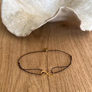Peut inclure: Un bracelet délicat composé d'un symbole infini doré centré sur un cordon noir fin. Le bracelet comprend une petite breloque en métal doré près du fermoir, présentant un design minimaliste avec une finition lisse et polie sur le symbole.