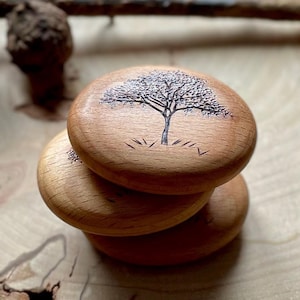 May include: Three wooden discs stacked on top of each other. The top disc has a simple line drawing of a tree with a few lines of grass at the bottom.