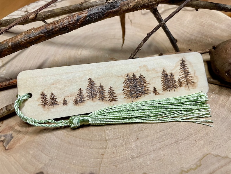 Handgebranntes Lesezeichen aus Ahorn mit Tannenwaldmotiv, mit ökologischem Klarlack versiegelt. Personalisierung auf der Rückseite möglich. Verschiedene Quasten und Bänder in verschiedenen Farben zur Auswahl.