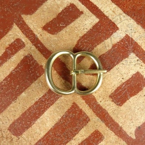 May include: A gold-coloured metal buckle with a smooth, rounded design. The buckle is approximately 5 cm in length and 2.5 cm in width.