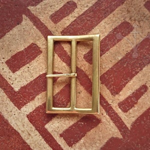 Puede incluir: Una hebilla de metal de color dorado con forma rectangular y dos barras horizontales. La hebilla está sobre una superficie con un patrón rojo y blanco.
