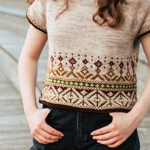 Op de afbeelding: Een vrouw draagt een beige gebreid topje met een geometrisch patroon in bruin, groen en rood. Het topje heeft korte mouwen en een ronde hals.