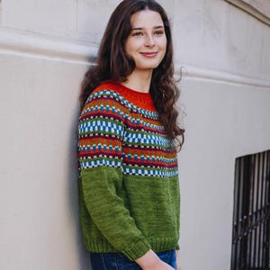 May include: A knitted jumper with a colour block design. The top section has a pattern of blue, red, and white geometric shapes on a black background. The sleeves and lower part are green. The person is wearing blue jeans and red trainers.
