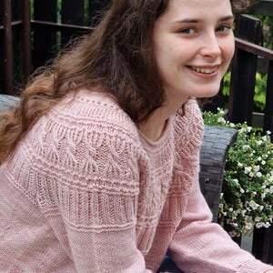 May include: A light pink, crew neck sweater with intricate cable knit and textured patterns. The long-sleeved garment features detailed stitching across the chest and sleeves. The sweater is made of a soft, possibly wool or cotton blend, material.