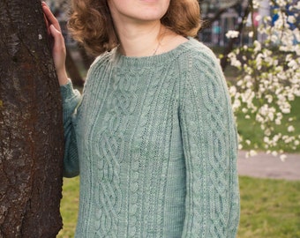 May include: A woman wearing a light green cable knit sweater with a loose fit. She is standing next to a tree with white blossoms.