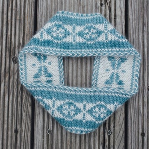 Op de afbeelding: Een gebreide col in een ruitvorm, met een herhalend patroon van witte en blauwgroene geometrische ontwerpen. De col is gemaakt van zacht garen en wordt gepresenteerd op een verweerd houten oppervlak. Het ontwerp bevat een vierkante opening.