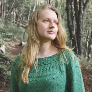 May include: A woman with long blonde hair wearing a green knitted sweater with a delicate pattern. She is standing in a forest with tall trees in the background.