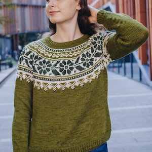 Può includere: Maglione verde oliva con motivo bianco e verde sullo sprone. Il maglione a maniche lunghe presenta uno scollo tondo e un motivo floreale dettagliato sul petto e sulle spalle. Il maglione è abbinato a jeans blu.