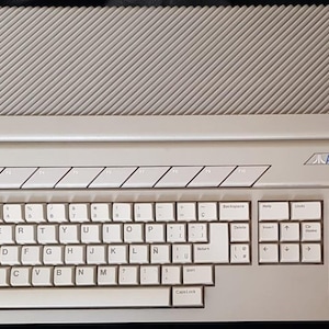 Peut inclure: Un ordinateur Atari 1040ST vintage, avec un clavier gris clair et une section supérieure assortie avec un design strié. Le clavier a des touches blanches avec des lettres noires. Le logo Atari et "1040ST" sont visibles.