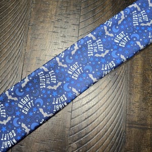 May include: A blue necktie with a pattern of stars, moons, and bats. The text "NIGHT SHIFT" and "SLEEP IS FOR SISSIES" is printed on the tie. The tie is made of a soft fabric.