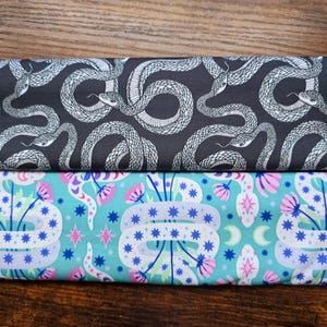 May include: Two fabric patterns are shown. The top pattern is dark grey with a white snake design. The bottom pattern is turquoise with a floral and snake design. The floral design includes pink flowers, blue stars, and white snakes.