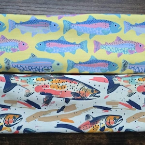 May include: Two fabric swatches with a repeating pattern of trout fish. The top swatch is yellow with blue and pink trout. The bottom swatch is white with orange, pink, and blue trout.