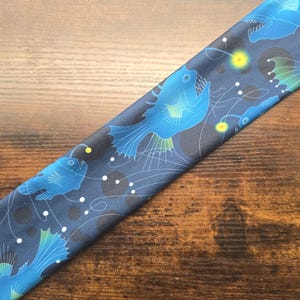 May include: A dark blue fabric with a repeating pattern of blue anglerfish, white dots, and yellow glowing orbs. The fabric is angled across a wooden surface.