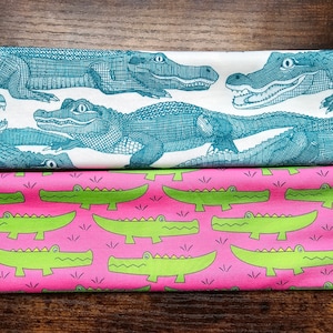 May include: Two fabric swatches with alligator designs. The top swatch features a white background with teal alligators. The bottom swatch features a pink background with green alligators.