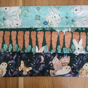 May include: Three fabric panels with different patterns. The top panel is light blue with white bunnies and green leaves. The middle panel is green with brown carrots and white bunnies. The bottom panel is dark green with white owls and purple flowers.