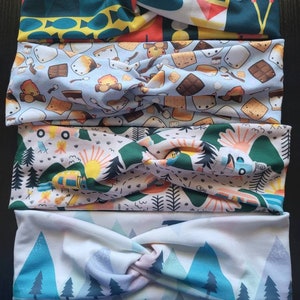 Peut inclure: Quatre bandeaux pour cheveux en tissu avec des motifs sur le thème du camping. Les motifs comprennent des montagnes, des feux de camp, des s'mores et des arbres de différentes couleurs. Les bandeaux sont torsadés à l'avant.