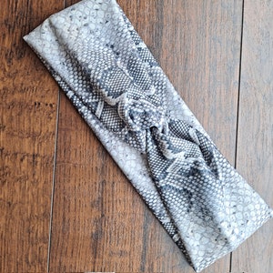 May include: A grey and white snakeskin print headband with a knotted design. The fabric has a textured appearance, and the headband is angled across a wooden surface.