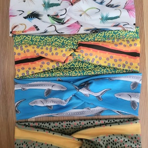 May include: Four different patterned headbands, each with a twist knot in the center. The top headband has a white background with a pattern of fish and fishing flies. The second headband has a yellow and green background with a geometric pattern. The third headband has a blue background with a pattern of sturgeon fish. The bottom headband has a green and yellow background with a pattern of brown spots.