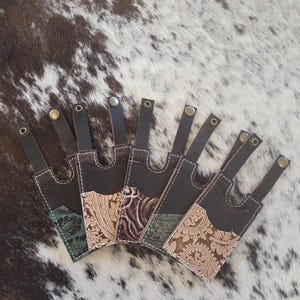 May include: A collection of leather knife sheaths with decorative embossed patterns in shades of brown, tan, and teal. Each sheath has a dark brown leather strap with a metal rivet.