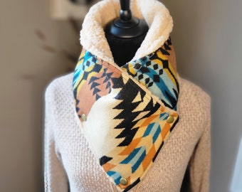 Puede incluir: Un calentador de cuello de forro polar con estampado blanco, negro, azul y naranja con forro de piel sintética. El calentador de cuello está doblado por la mitad y tiene un cierre a presión.