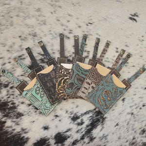 May include: A collection of leather cardholders in various colors and textures. Each cardholder features a unique embossed design, including floral and geometric patterns. The cardholders have a brown leather strap and a snap closure.