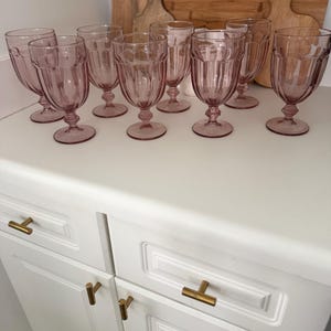 May include: A set of ten pink-tinted, stemmed water goblets with vertical ribbed detailing. The glasses are arranged on a white countertop, with a white cabinet and gold-toned hardware in the foreground. Wooden cutting boards are in the background.