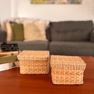 May include: Two square woven storage boxes with lids. The boxes are made of a light brown, natural fiber material and sit on a dark brown wooden table. The boxes are empty and ready to store small items. The background includes a sofa and artwork.