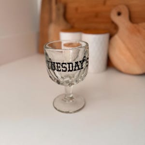 May include: Clear glass goblet with a textured bowl and a round base. The word "TUESDAY'S" is printed in black on the glass. The goblet is on a white surface.