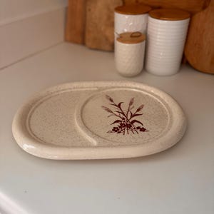May include: An oval-shaped, off-white ceramic serving platter with a speckled surface. The platter features a floral design in maroon on one side and a curved indentation. Kitchenware.