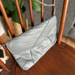 May include: A gray leather clutch purse with a flap closure and a long strap. The purse is sitting on a wooden chair.