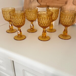 May include: A set of six amber-colored glass goblets, each featuring a textured, repeating oval pattern on the bowl, a short stem, and a circular base. The glassware is arranged in two rows, showcasing a consistent warm, translucent orange hue.