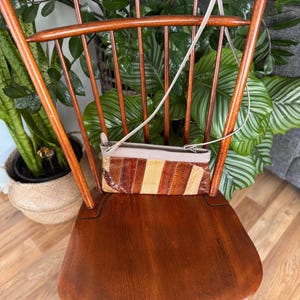 May include: A vintage wooden chair with a high back and a small, rectangular purse. The purse has a beige top and a patterned front with brown, tan, and orange stripes. The chair is made of dark wood and is surrounded by green plants.