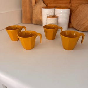 May include: Four mustard-yellow, faceted cups with angular handles are arranged on a white surface. The cups have a geometric design and are empty. In the background are wooden cutting boards and white ceramic canisters with wooden lids.