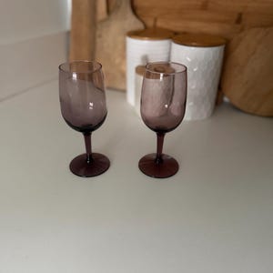 May include: Two wine glasses made of translucent purple glass. Each glass features a rounded bowl, a slender stem, and a circular base. The glass material has a smooth, polished finish, and the color is consistent throughout the bowl, stem, and base.