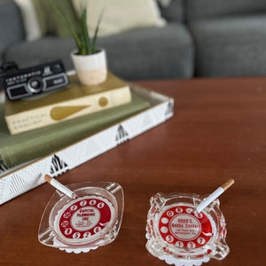 May include: Two vintage glass ashtrays with red and white phone dial designs. One ashtray has the text "CAPITOL PLUMBING INC.  RUBBER HOSE SPECIALISTS" and the other has the text "ODDO'S Hobby Center 238 Shaw Ave. McKeesport, Pa."